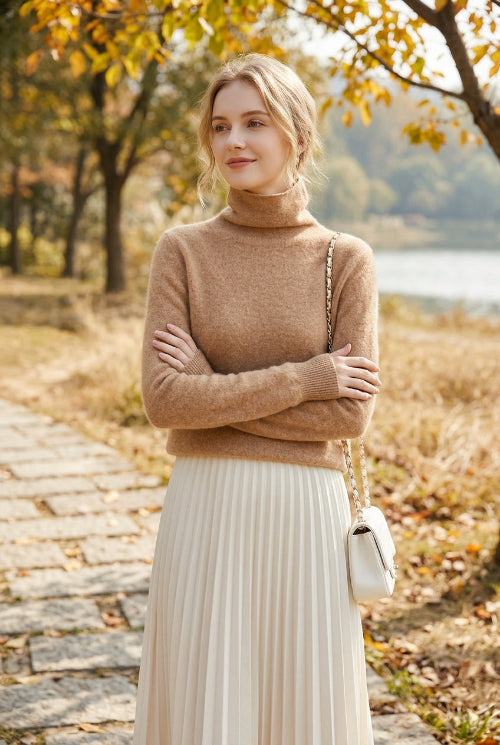 A person wearing a fuzzy, cropped turtleneck sweater in a warm camel beige color, paired with a light-colored skirt and holding a white shoulder bag with a silver chain strap.