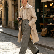 A man walking on a cobblestone street wearing a beige trench coat, a cream shirt, and pinstripe trousers, holding a camera. A cafe is visible in the background.
