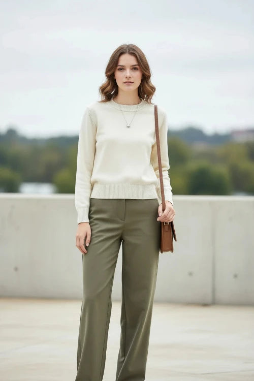 A woman wearing a cream-colored crewneck knit sweater, black sunglasses, and a small silver pendant necklace, with a brown shoulder bag strap visible over her shoulder.