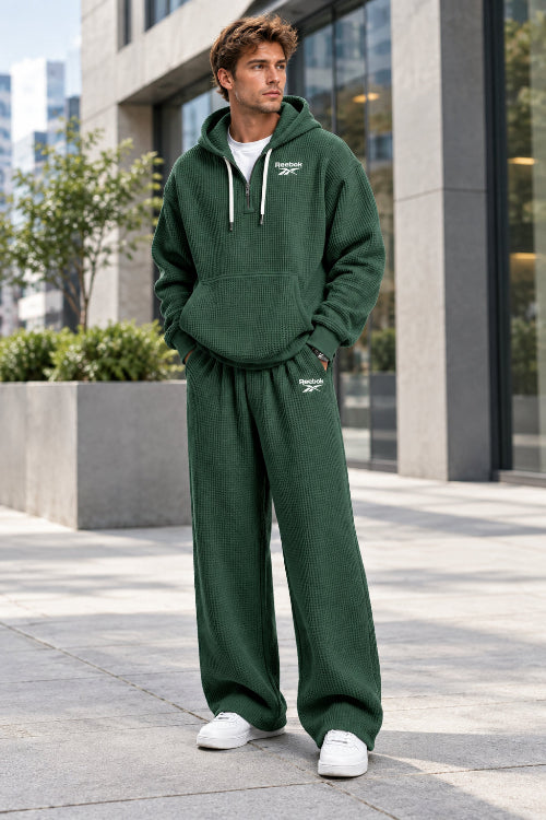 A person wearing a dark green waffle-knit hoodie and matching sweatpants set with small white Reebok logos, shown 