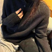 A person wearing a dark charcoal gray button-up knit cardigan sweater, sitting on a couch with a gray bag and light gray pants visible.