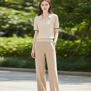 A woman wearing a beige short-sleeve polo shirt with black trim on the sleeve cuffs, paired with matching beige pants, set against a soft neutral indoor background.