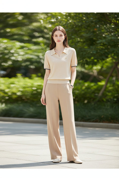 A woman wearing a beige short-sleeve polo shirt with black trim on the sleeve cuffs, paired with matching beige pants, set against a soft neutral indoor background.