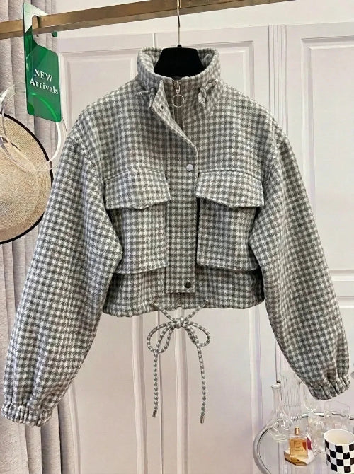 Gray and white checked utility jacket on a hanger, showing a raised collar, ring-pull zipper, chest flap pockets, front snaps, elasticated cuffs and a tied drawcord hem