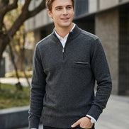 A man wearing a dark gray quarter-zip knit sweater over a white collared shirt and dark jeans, standing outdoors in front of a modern building.