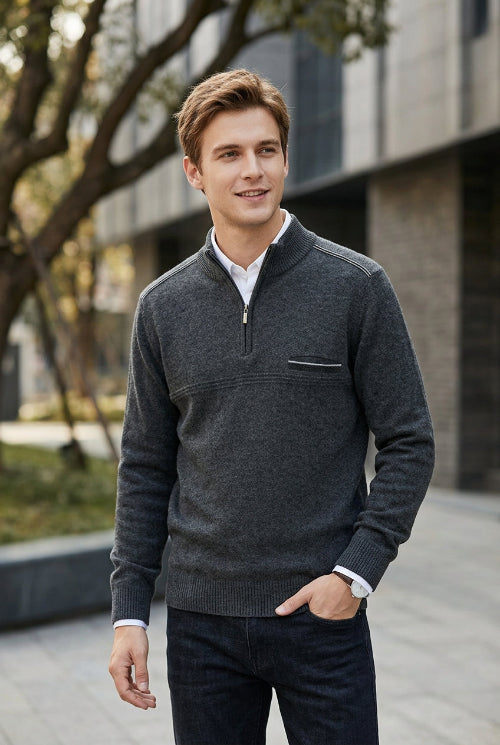 A man wearing a dark gray quarter-zip knit sweater over a white collared shirt and dark jeans, standing outdoors in front of a modern building.