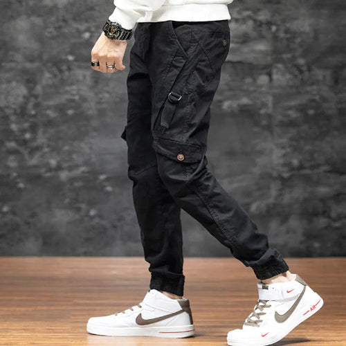 Side view of a model wearing black cargo pants with a buttoned thigh pocket, side strap, and gathered ankle cuffs, paired with white high-top sneakers.