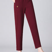 A model wearing high-waisted, straight-leg burgundy trousers with a small gold embroidered bird logo on the left thigh, paired with a black long-sleeve top and black pointed-toe heels against a dark gray studio backdrop.