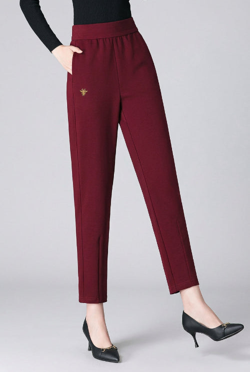 A model wearing high-waisted, straight-leg burgundy trousers with a small gold embroidered bird logo on the left thigh, paired with a black long-sleeve top and black pointed-toe heels against a dark gray studio backdrop.