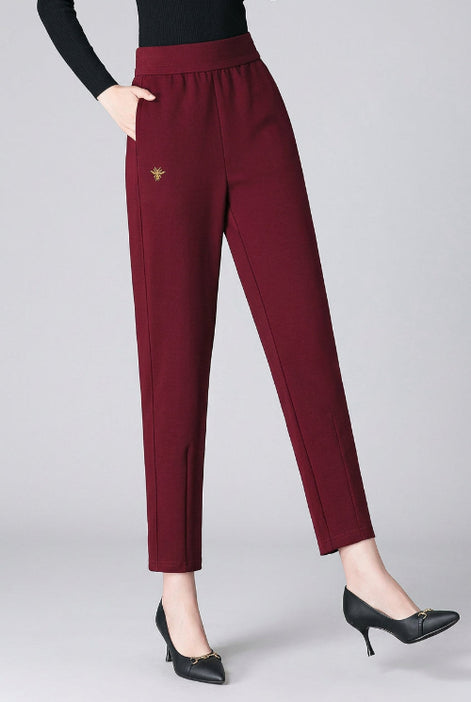 A model wearing high-waisted, straight-leg burgundy trousers with a small gold embroidered bird logo on the left thigh, paired with a black long-sleeve top and black pointed-toe heels against a dark gray studio backdrop.