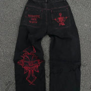 Back view of black wide-leg jeans laid flat on a gray surface, featuring purple lettering and a skeleton graphic on the back pockets and a large purple cross-style design on one leg.