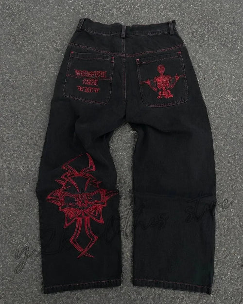 Back view of black wide-leg jeans laid flat on a gray surface, featuring purple lettering and a skeleton graphic on the back pockets and a large purple cross-style design on one leg.