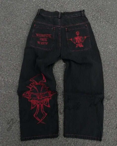 Back view of black wide-leg jeans laid flat on a gray surface, featuring purple lettering and a skeleton graphic on the back pockets and a large purple cross-style design on one leg.