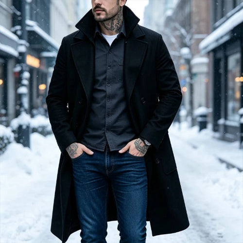 A man wearing a long black wool-blend overcoat over a dark button-up shirt and blue jeans, standing on a snow-covered city street 