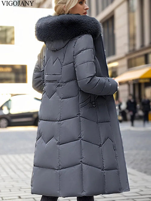 A woman wearing a long gray puffer coat with a large dark faux-fur collar, photographed outdoors on a city street. The coat has horizontal quilting and a hood.