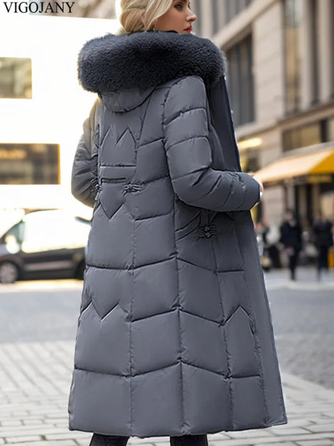 A woman wearing a long gray puffer coat with a large dark faux-fur collar, photographed outdoors on a city street. The coat has horizontal quilting and a hood.