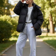 Person wearing a black puffer jacket and white pants against a gray wall.