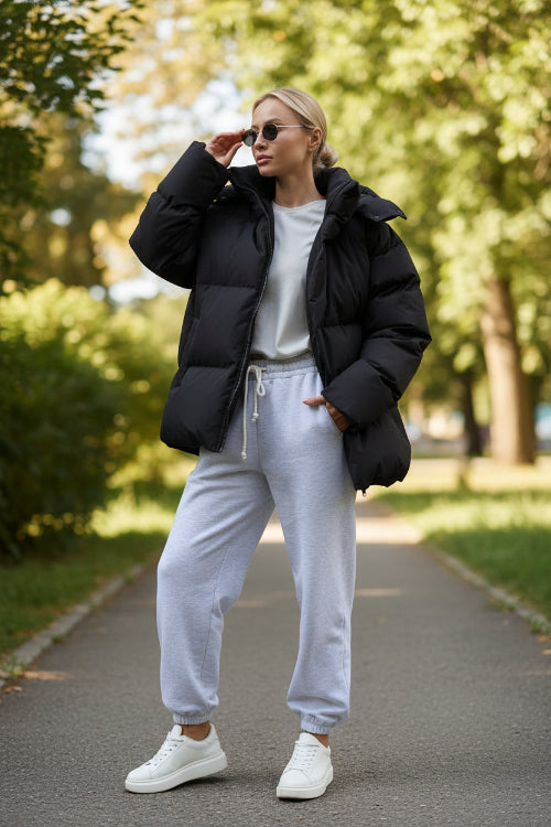 Person wearing a black puffer jacket and white pants against a gray wall.