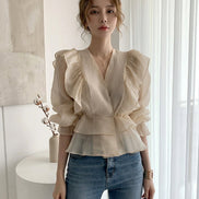 A woman wearing a beige ruffled blouse with a V-neckline and puffed sleeves, paired with blue jeans. The brand name 'Yukiesue' is visible in the top left corner.