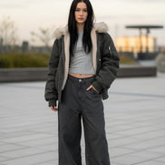 Person wearing a long dark coat with fur trim, standing on a wooden deck.