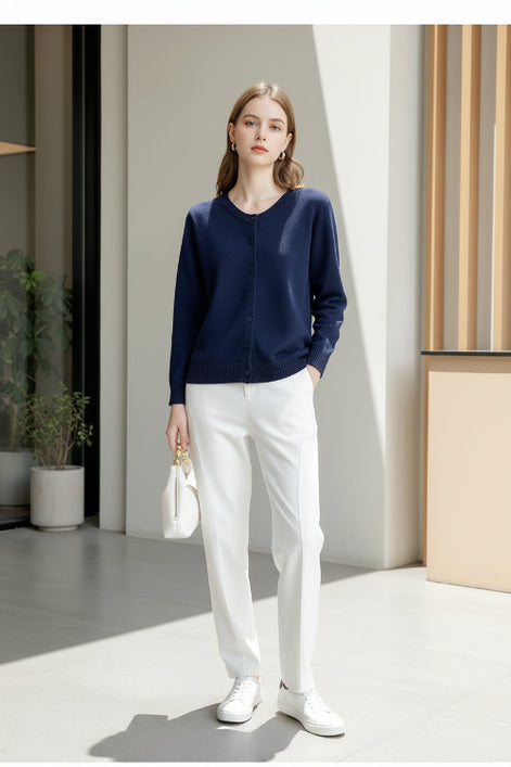 A woman wearing a navy blue long-sleeve button-up knit sweater and white pants, holding a white handbag 