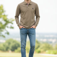 Beige polo shirt with a brand logo held by a hand, with a blurred background of books and shirts.