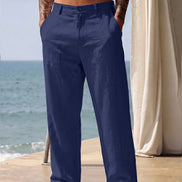 A man wearing navy blue linen trousers stands barefoot on a patio with the ocean and a cream curtain in the background.