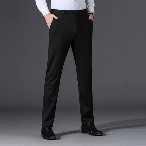 A man wearing slim-fit black dress pants with a white dress shirt and black dress shoes, standing against a dark gray studio background.