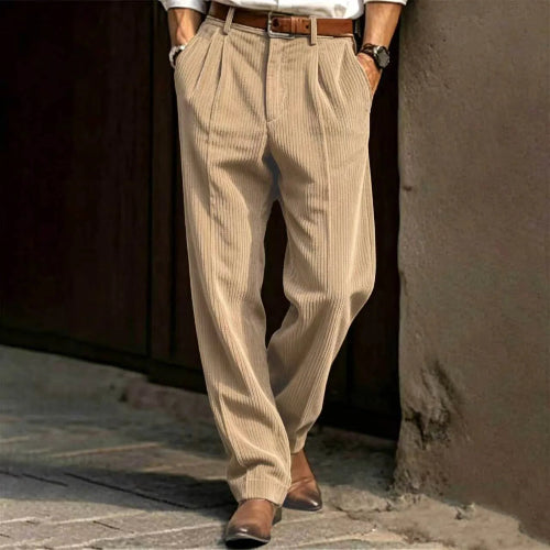Person wearing beige pleated pants and brown shoes against a textured wall.