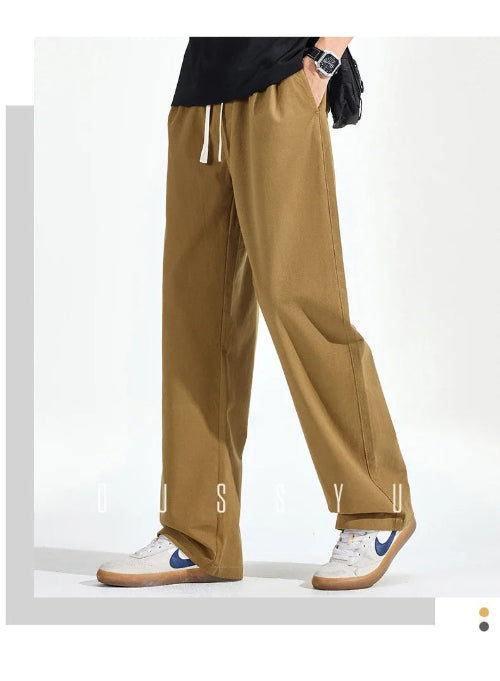 Model wearing khaki loose-fit straight-leg pants with a white drawstring and one hand in a side pocket, paired with white sneakers.