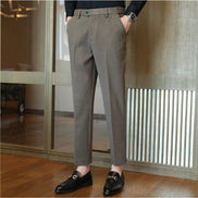 A person wearing slim-fit taupe gray trousers, paired with a black long-sleeve shirt and black loafers, standing in an indoor setting with wooden floors and glass doors.