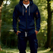 A man wearing a navy blue zip-up hoodie and matching cargo pants stands in a sunlit forest. The hoodie has gray drawstrings with orange tips, 