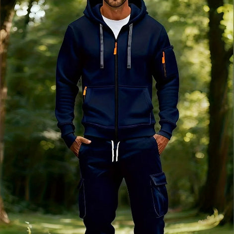 A man wearing a navy blue zip-up hoodie and matching cargo pants stands in a sunlit forest. The hoodie has gray drawstrings with orange tips, 