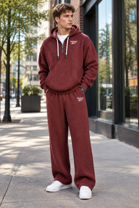 A man wearing a matching Reebok waffle-knit hoodie and sweatpants set in a dusty burgundy red color, shown against a plain white background.