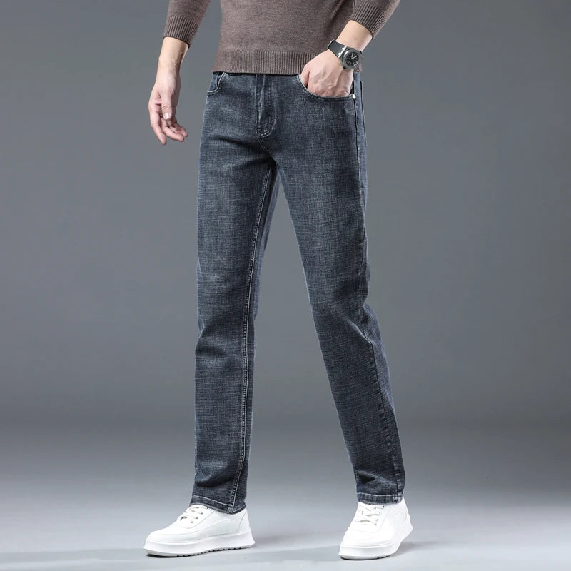 A person modeling slim-fit dark gray denim jeans with a brown sweater and white sneakers against a gray studio background.