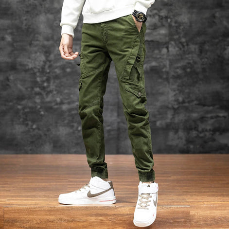 Model wearing olive green cargo pants with angled thigh pockets and gathered ankle cuffs, paired with white high-top sneakers against a dark wall.