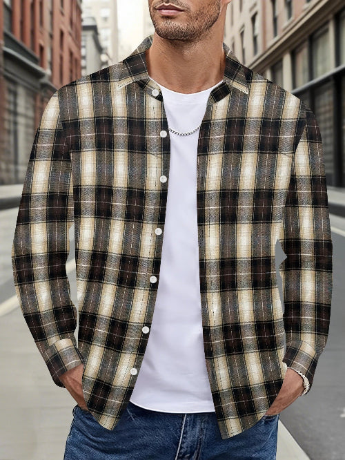 A man wearing a long-sleeve button-up plaid flannel shirt in black, beige, and brown over a white t-shirt and blue jeans, standing on a city street.