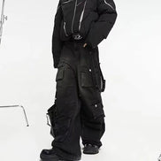 Model wearing washed black baggy cargo jeans with multiple flap pockets and hanging side straps, paired with a black padded jacket and chunky black shoes.