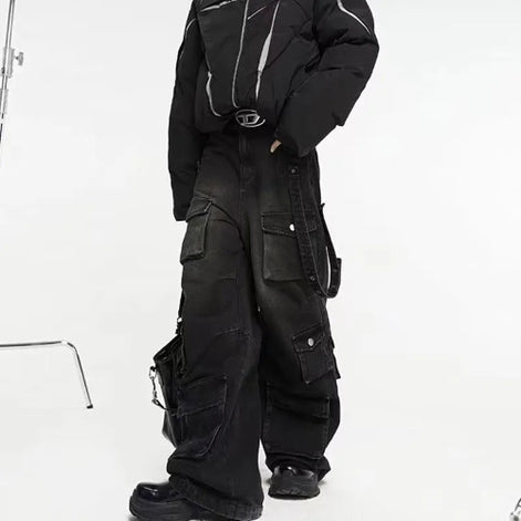 Model wearing washed black baggy cargo jeans with multiple flap pockets and hanging side straps, paired with a black padded jacket and chunky black shoes.