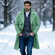 A man wearing a light green double-breasted overcoat over a dark gray shirt, white t-shirt, and blue jeans, standing in a snowy park.