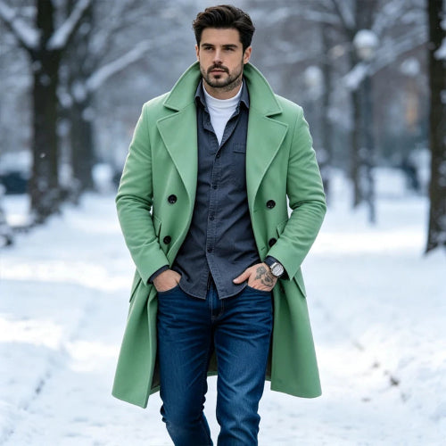 A man wearing a light green double-breasted overcoat over a dark gray shirt, white t-shirt, and blue jeans, standing in a snowy park.