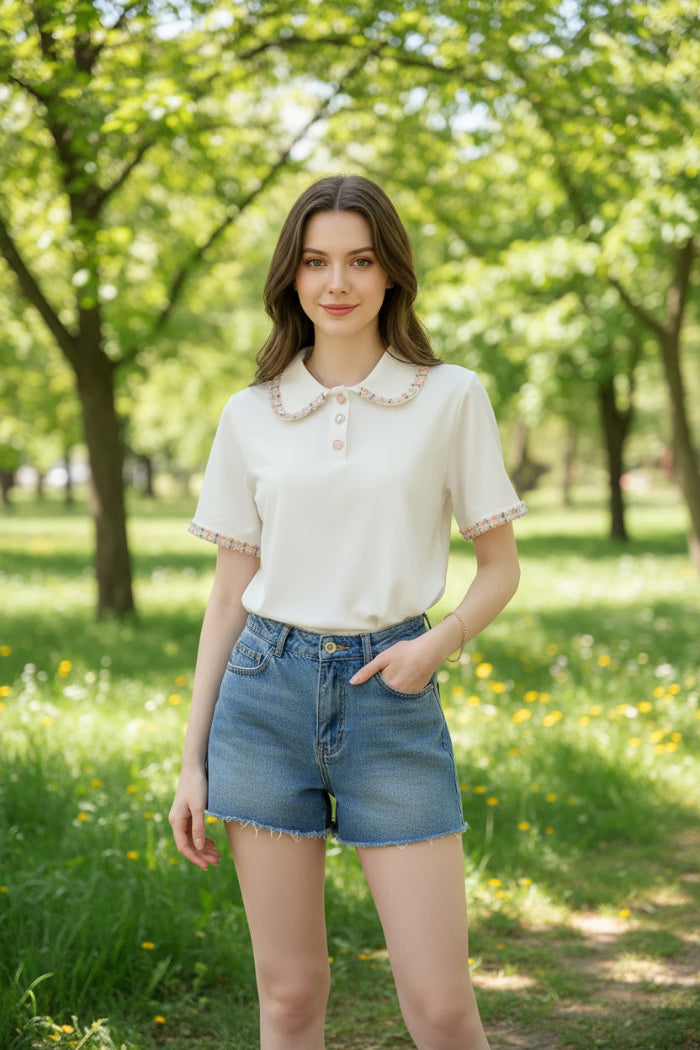 A woman wearing a short-sleeve cream-colored blouse with a decorative fringed Peter Pan collar and matching fringed sleeve cuffs, paired with denim shorts, sitting on a dark textured couch.