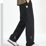 Side view of a model wearing black loose-fit straight-leg pants with a small sewn-on label and one hand in a pocket, paired with white sneakers.