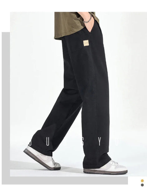 Side view of a model wearing black loose-fit straight-leg pants with a small sewn-on label and one hand in a pocket, paired with white sneakers.