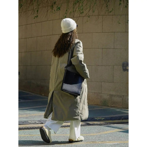 Rear view of a woman walking in an army green long padded coat with a center back vent, styled with white trousers and a black shoulder bag