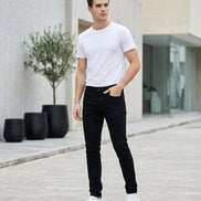 A person modeling slim black pants with a white t-shirt and white sneakers, standing in a retail store setting next to a white and yellow robot toy.