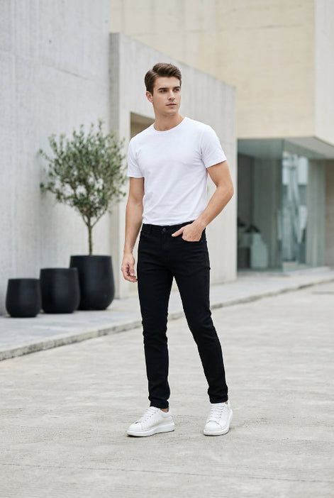 A person modeling slim black pants with a white t-shirt and white sneakers, standing in a retail store setting next to a white and yellow robot toy.