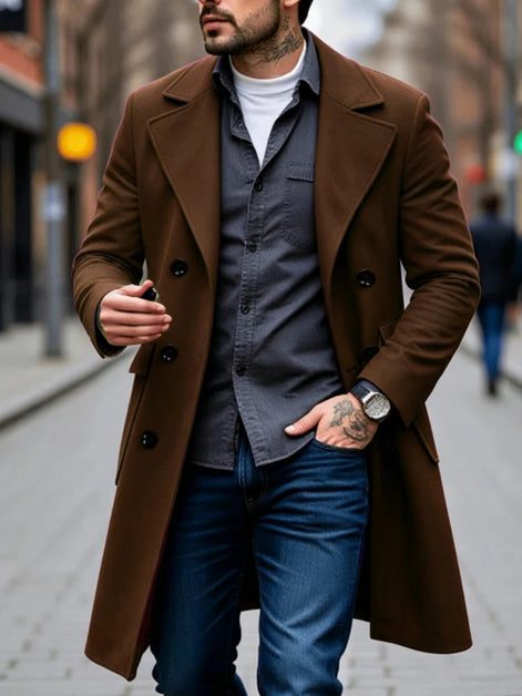 A man wearing a mid-length brown double-breasted overcoat over a dark gray button-up shirt, blue jeans, and a white t-shirt, walking on a city street.
