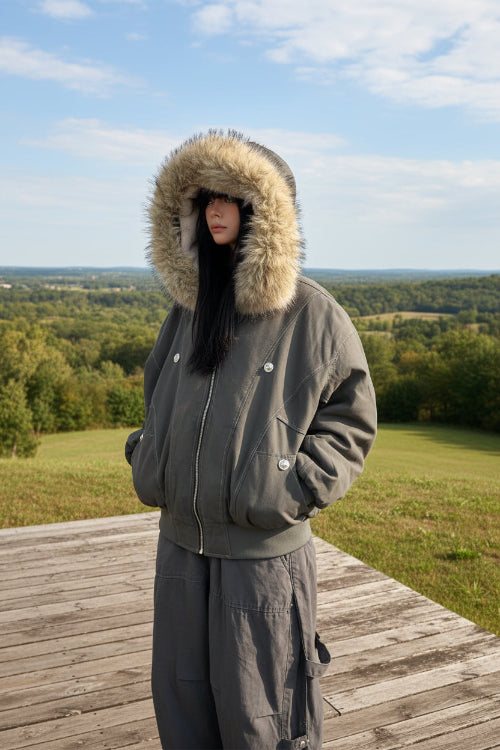 Person wearing a gray parka with a fur-lined hood against a white wall.