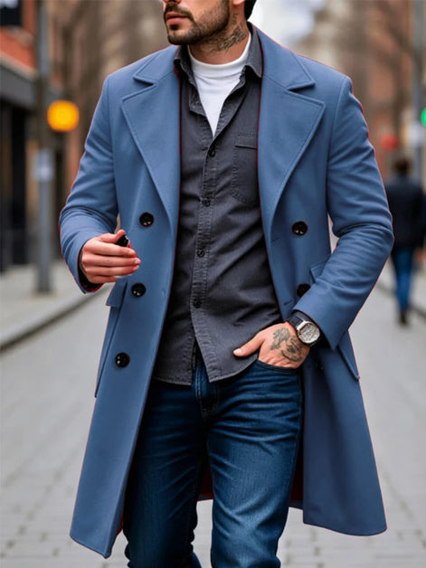 A man wearing a long blue double-breasted overcoat over a dark gray button-up shirt, white t-shirt, and blue jeans, walking on a blurred city street.